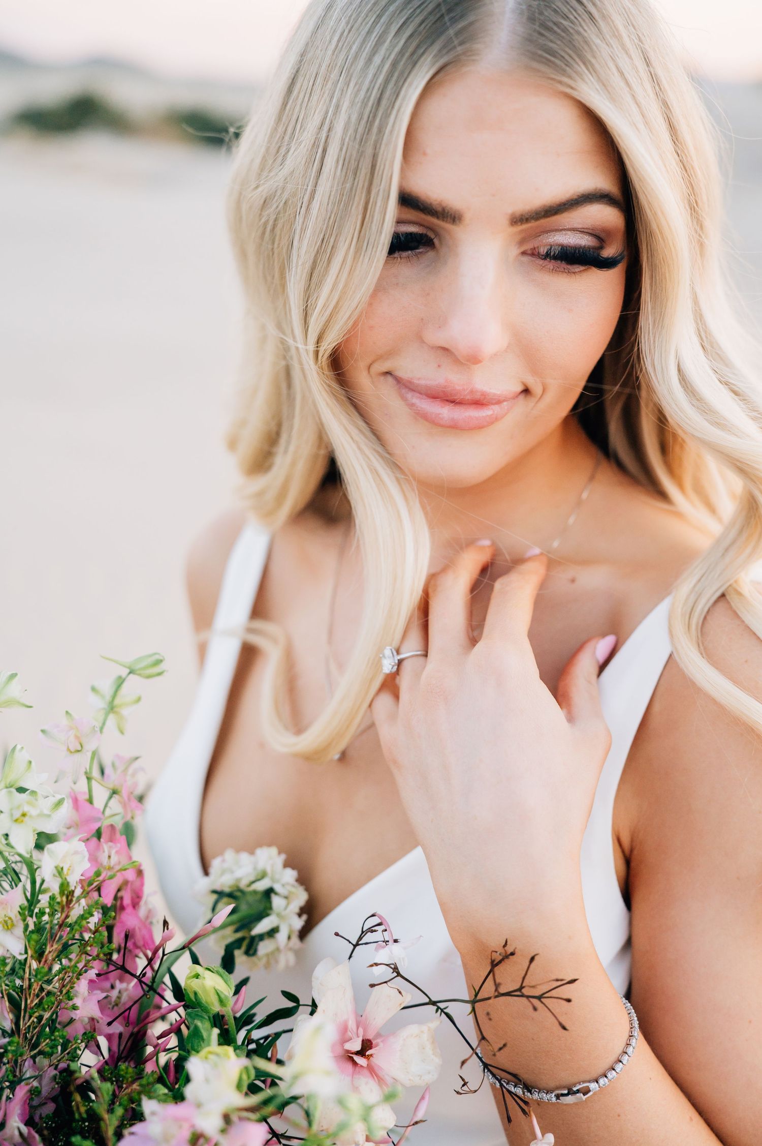 Delicate bridal portrait with blonde waves, natural makeup, white dress and pink wildflower bouquet against light background.