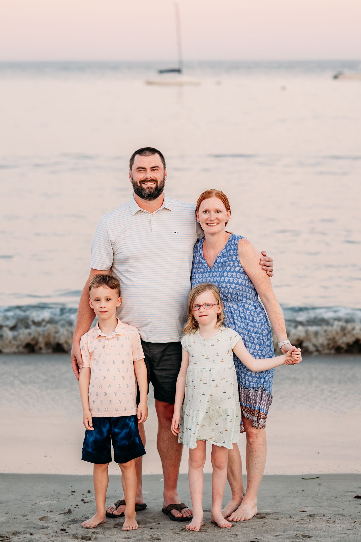 Family standing together on a beach at sunset with sailboats visible in the background across calm waters.