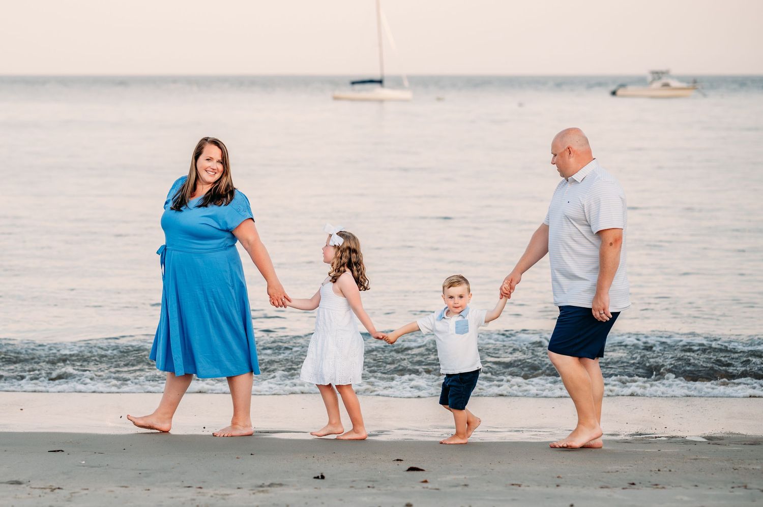 A family walks hand in hand along a peaceful beach at sunset with boats visible in the distance on the water.