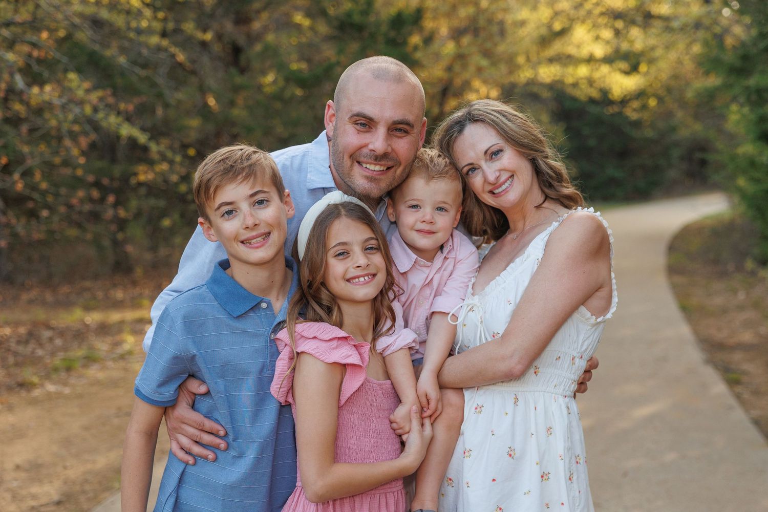 A loving family of five poses together on a dirt path surrounded by autumn foliage during golden hour.