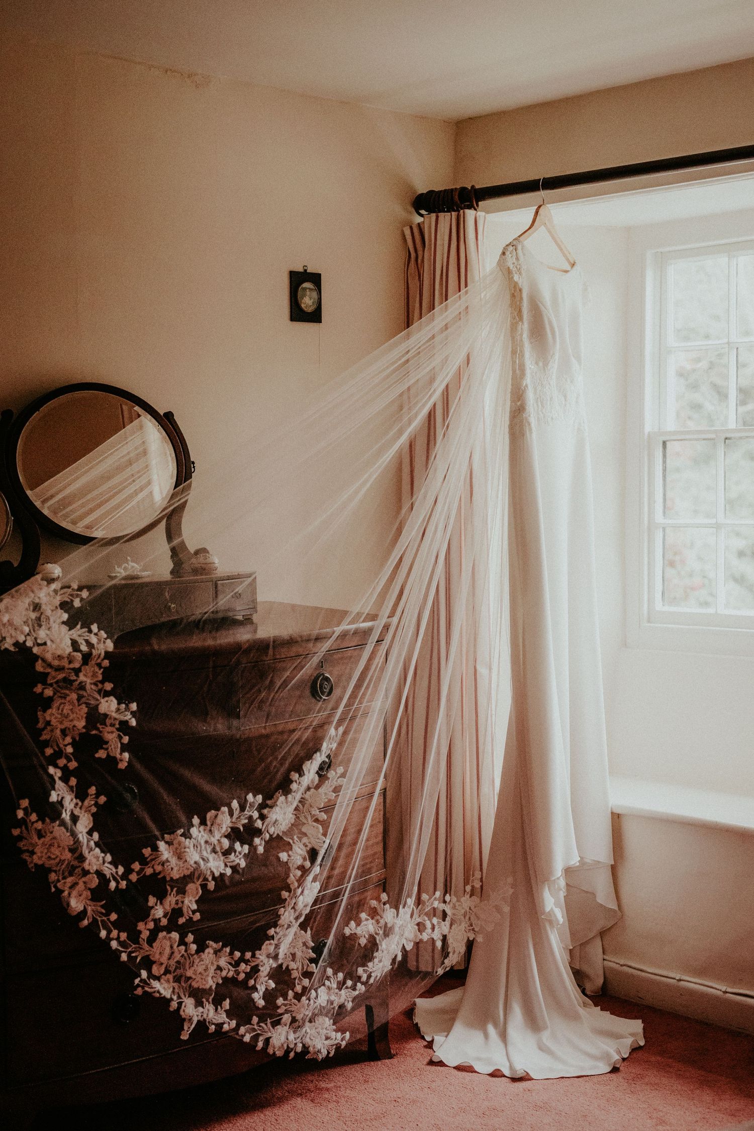 A wedding dress and veil hang elegantly by a window with a vintage piano draped in delicate floral decorations.