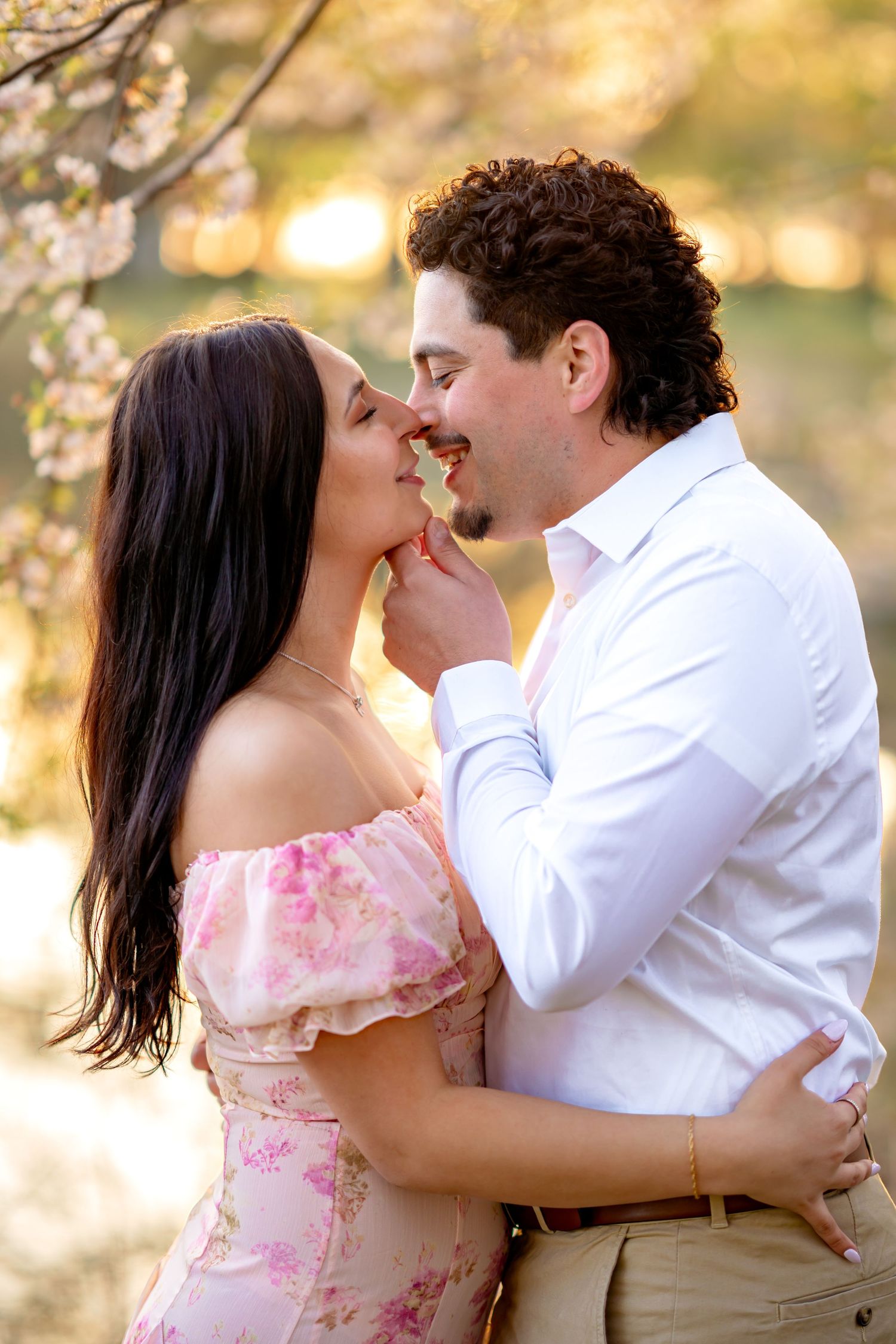 A romantic spring engagement photo of a couple embracing under cherry blossoms at sunset.