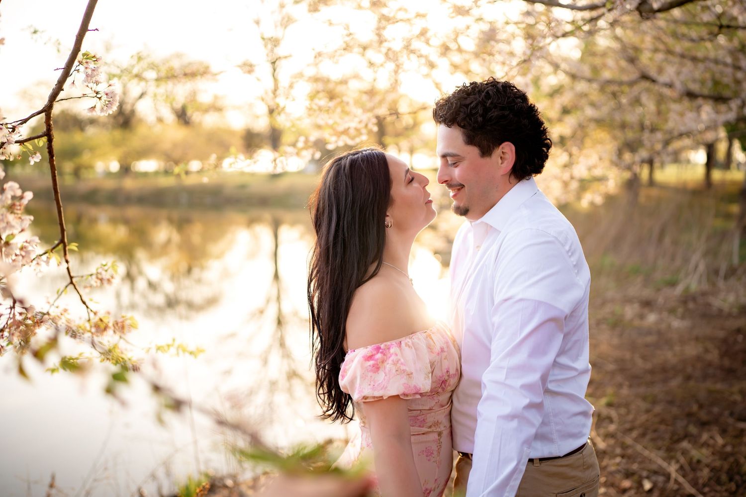 A romantic moment between a couple in springtime by a tranquil pond with blooming cherry blossoms above.