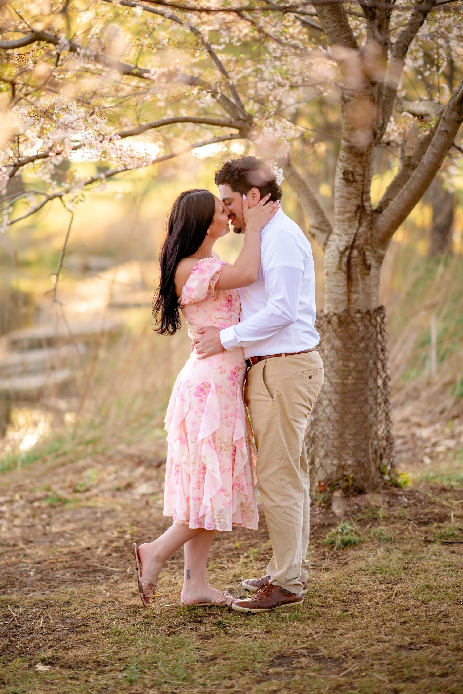 A romantic couple embraces under a blooming tree during golden hour, with her wearing a pink floral dress and him in khakis.