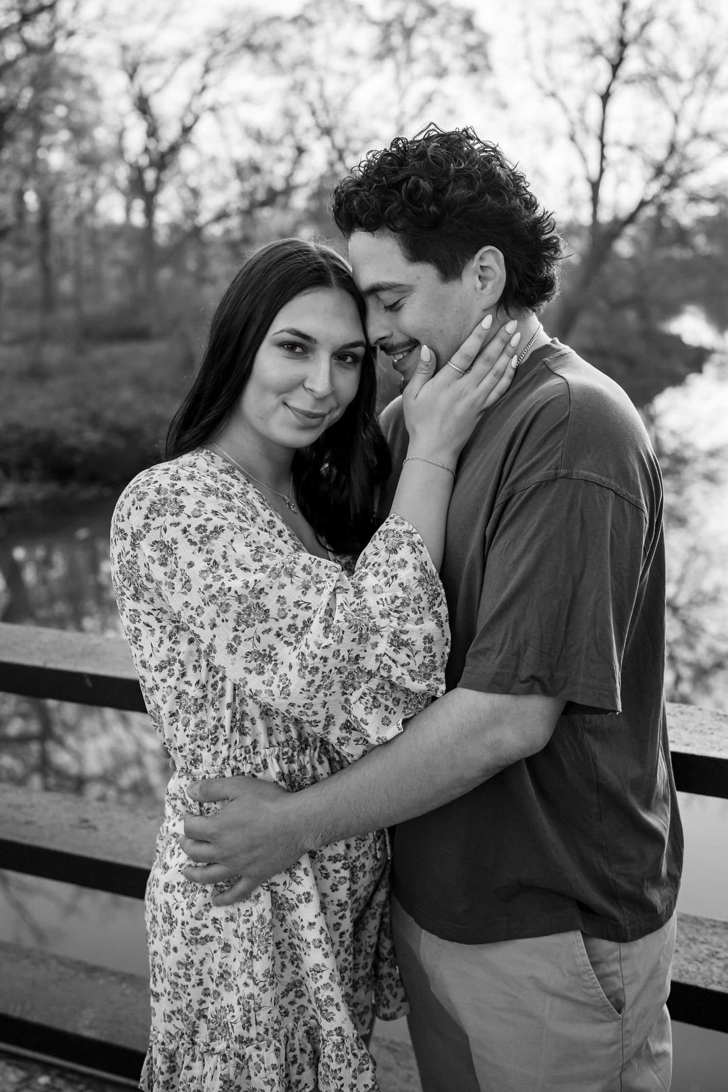 A couple shares an intimate moment in black and white while embracing on a bridge with trees in the background.