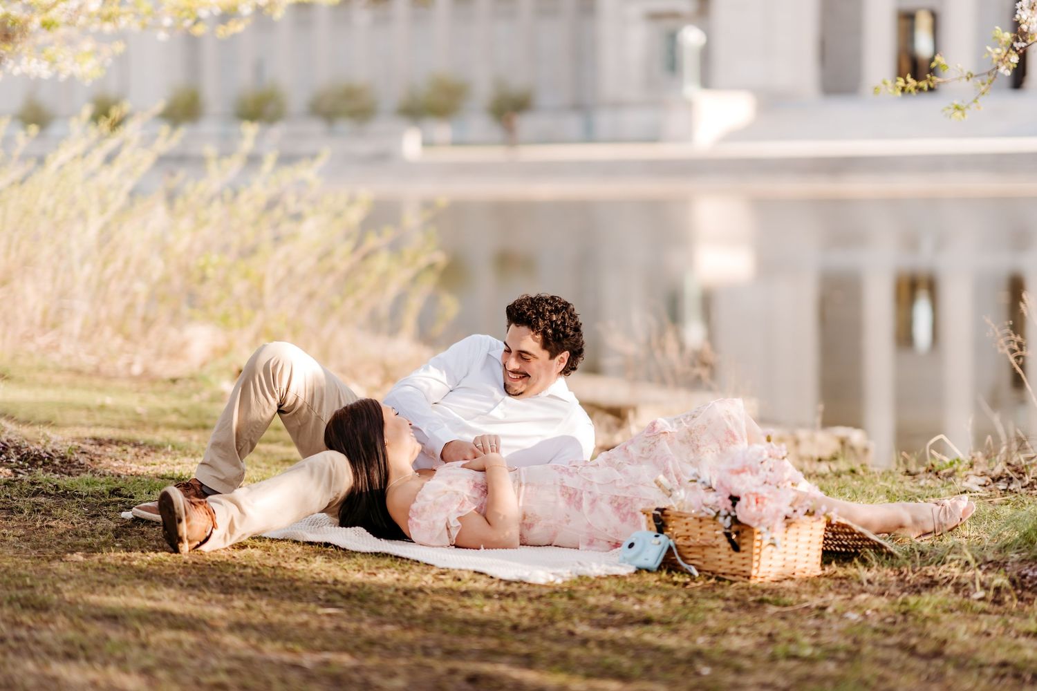 A couple in light colored outfits having a romantic picnic by a lakeside on a sunny afternoon.