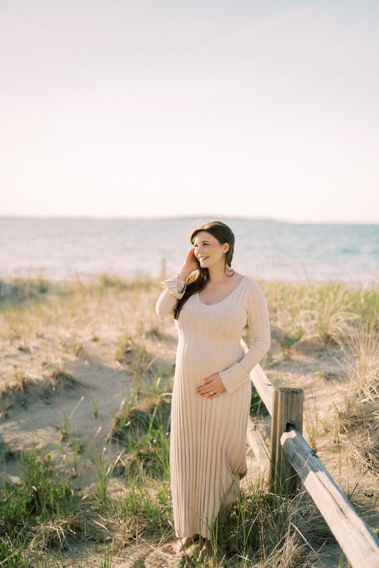 Woman in a cream long-sleeved maxi dress stands on a sandy beach dune during golden hour at sunset.