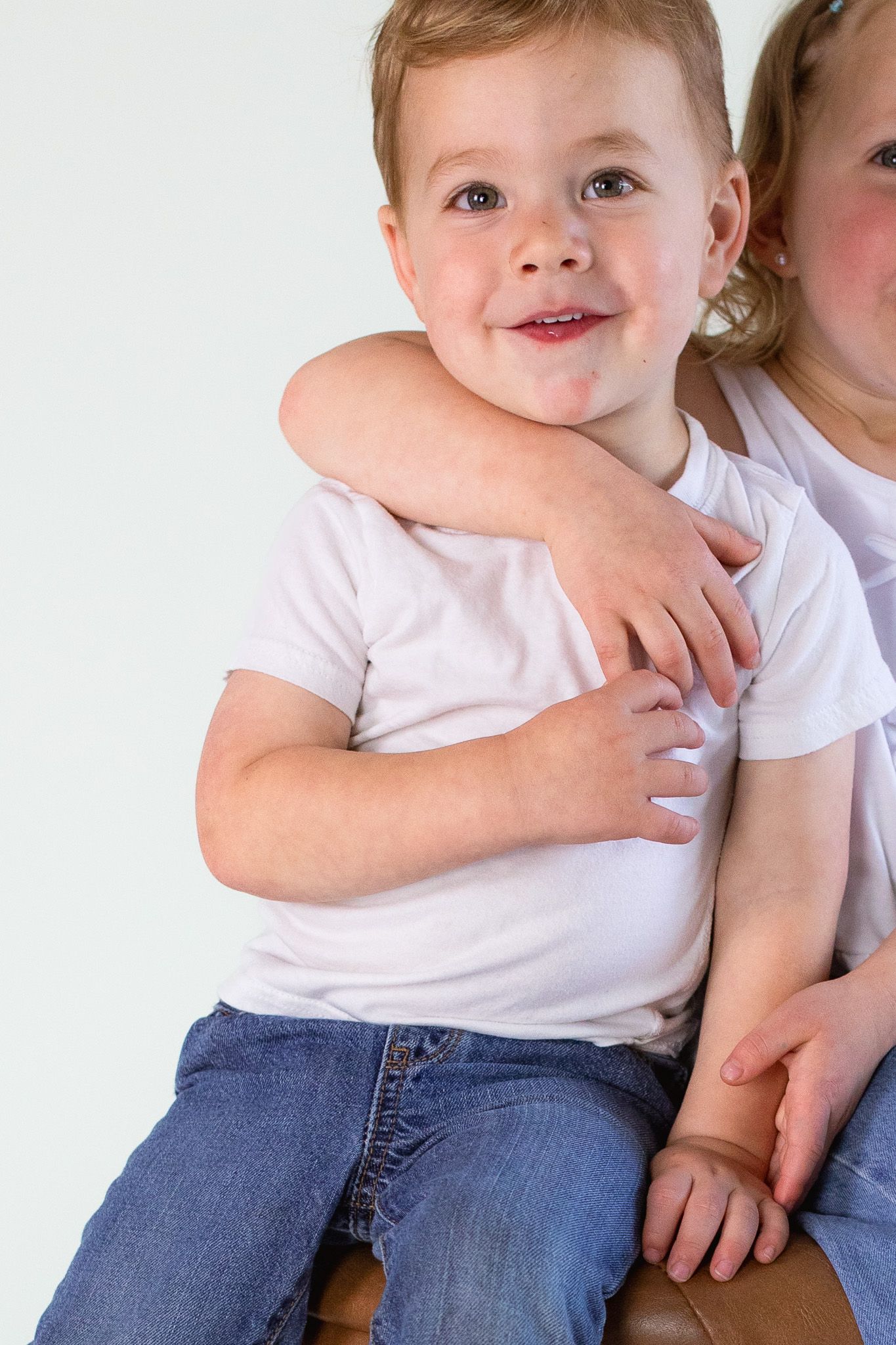 Child wearing a white t-shirt and jeans sitting close with someone else against white background.