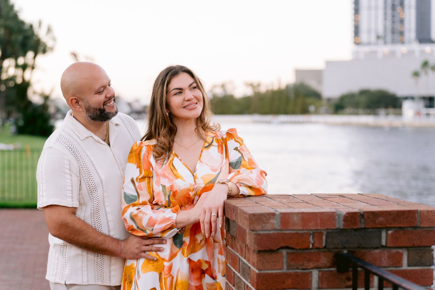 A couple in summer attire shares a tender moment by a brick wall overlooking the Hillsborough River at sunset.
