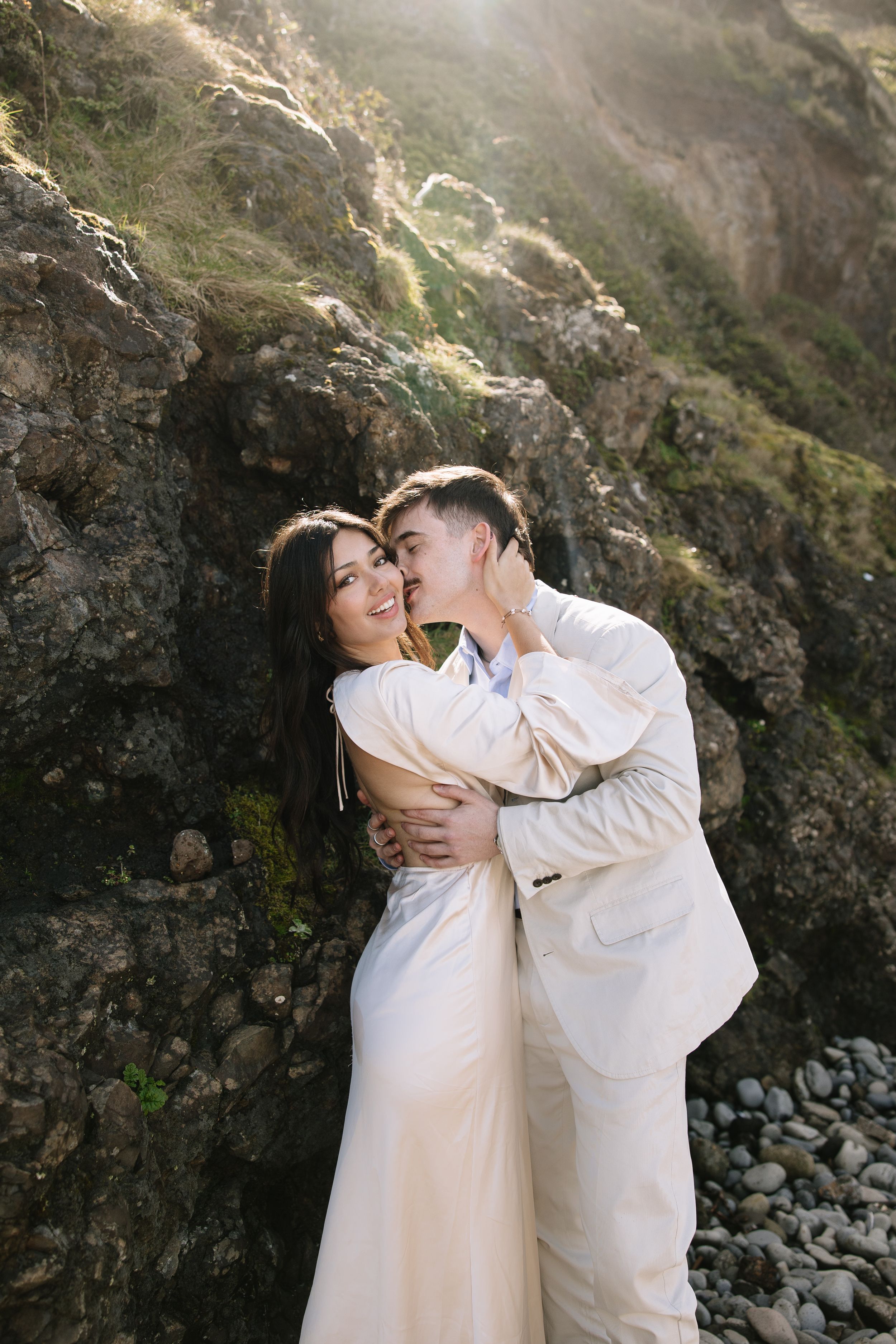 A romantic couple in white attire embrace against a dramatic rocky mountain backdrop during a sunlit photo session.