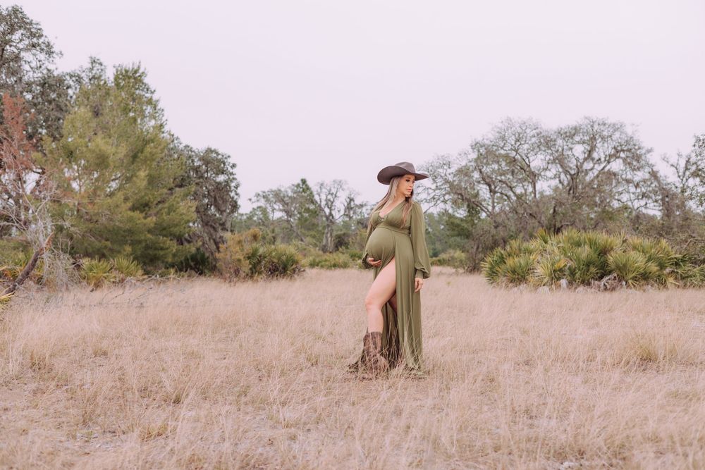 A person in a long olive dress and cowboy hat poses in a dry grass field surrounded by trees at golden hour.