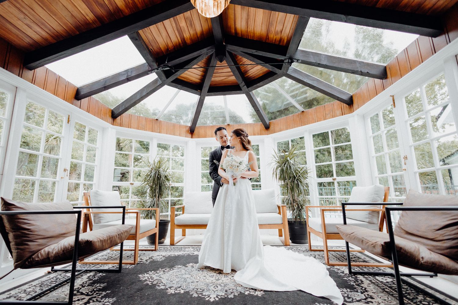 A romantic couple stands in an elegant greenhouse-style sunroom captured by Apertured Media Auckland Wedding Photographer.