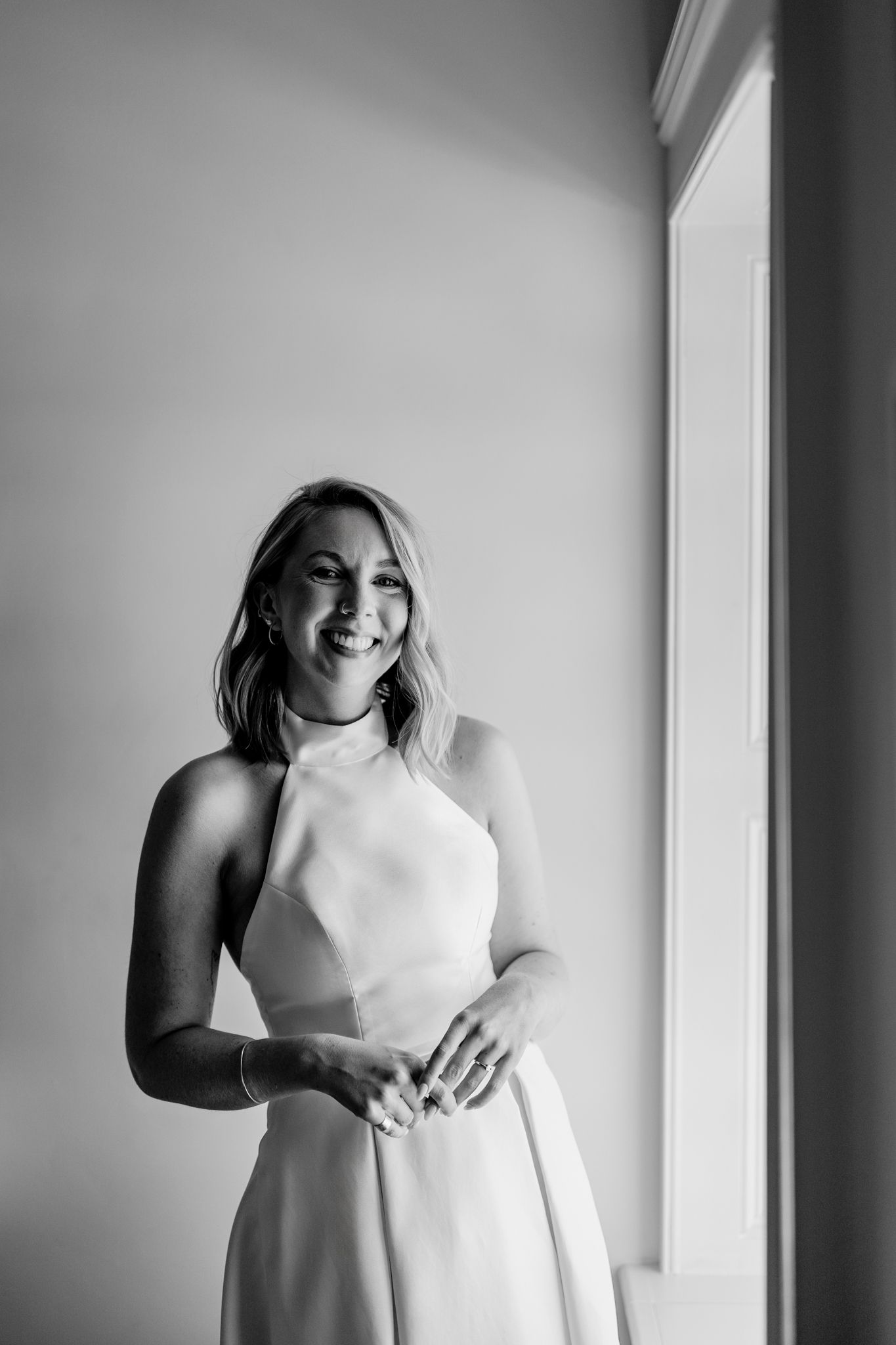 Black and white portrait of person in elegant white high-neck dress standing by window with natural light.