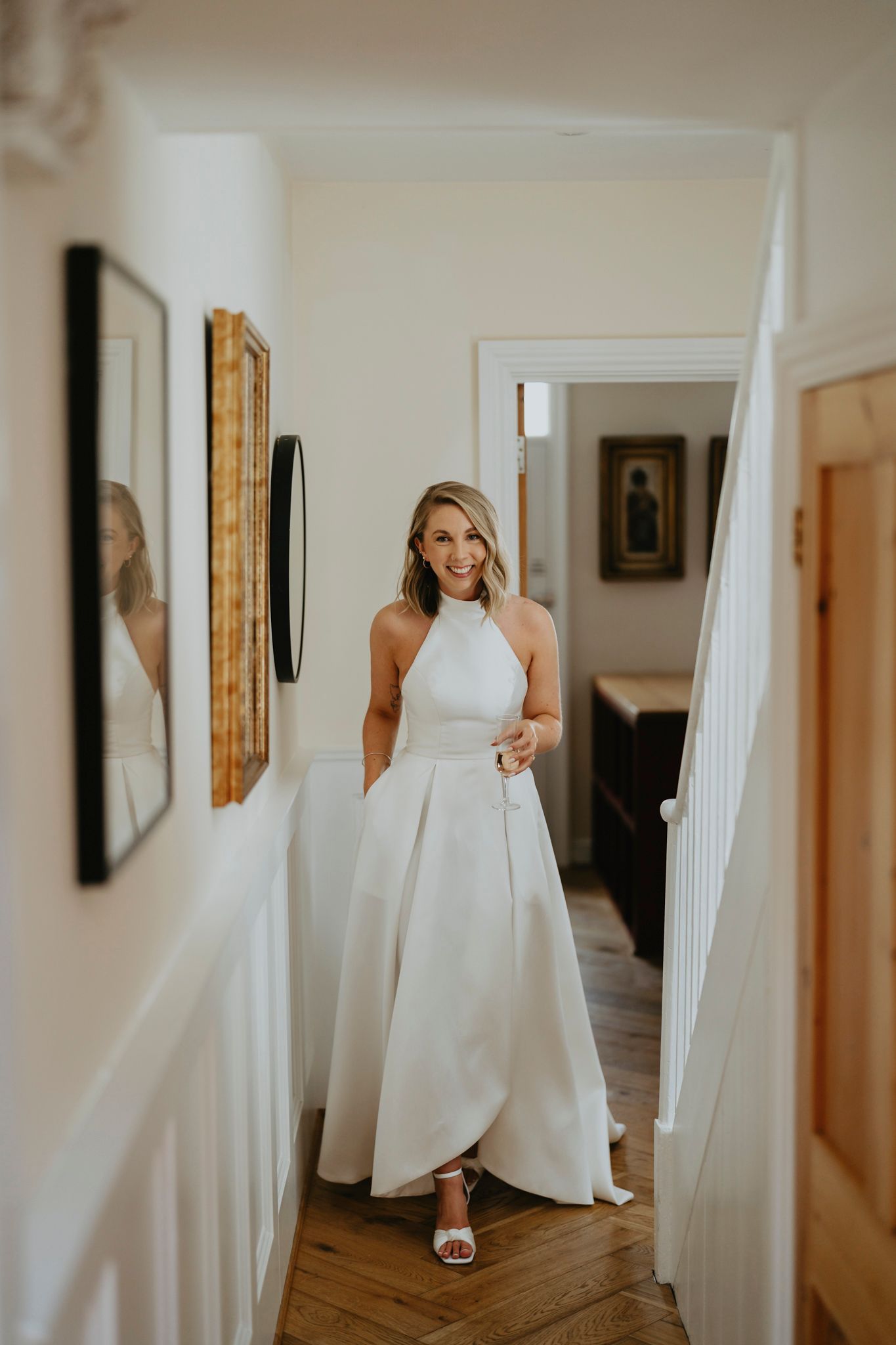 Person wearing an elegant white halter wedding dress stands in a hallway with hardwood floors and framed artwork on walls.