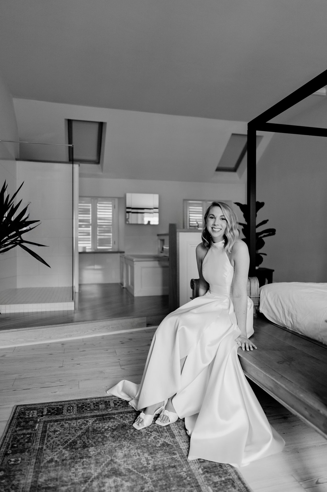 A bride in an elegant white wedding gown sits on a modern bed in a minimalist room with hardwood floors.