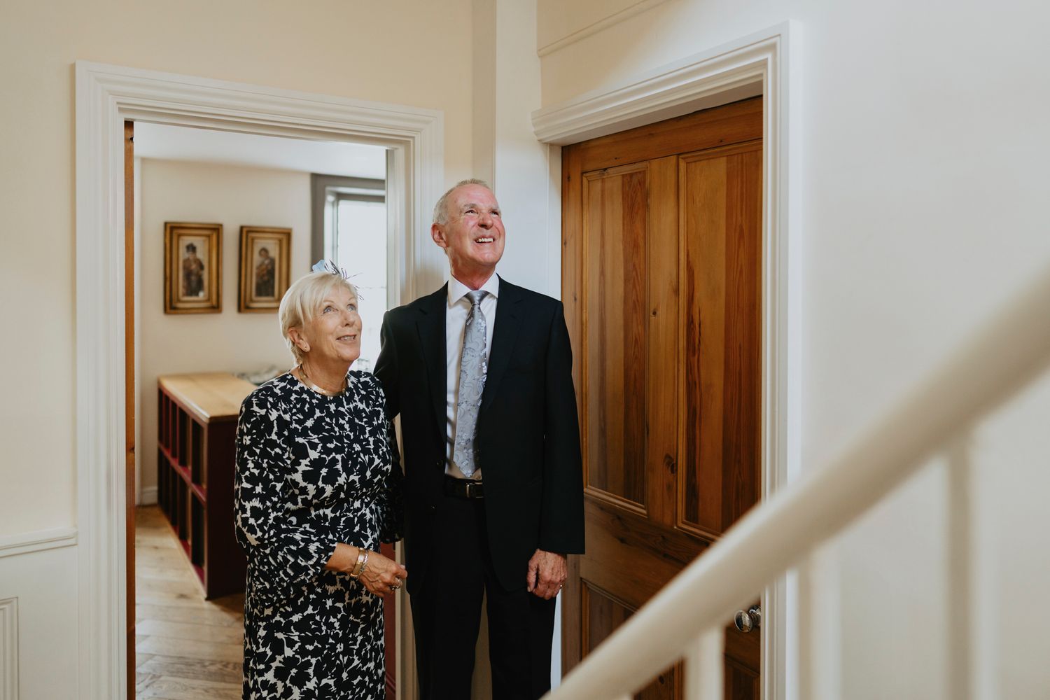 An elderly couple stands together in a hallway near a wooden door and staircase in a warmly lit interior.