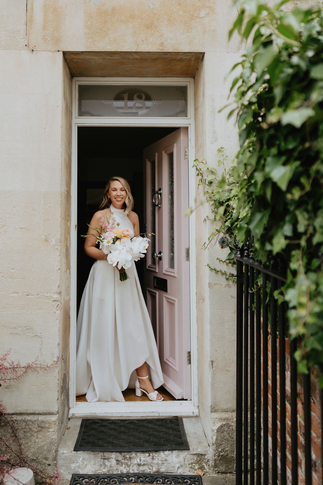 A bride holding a white bouquet stands in a pink doorway entrance with climbing vines framing the scene.