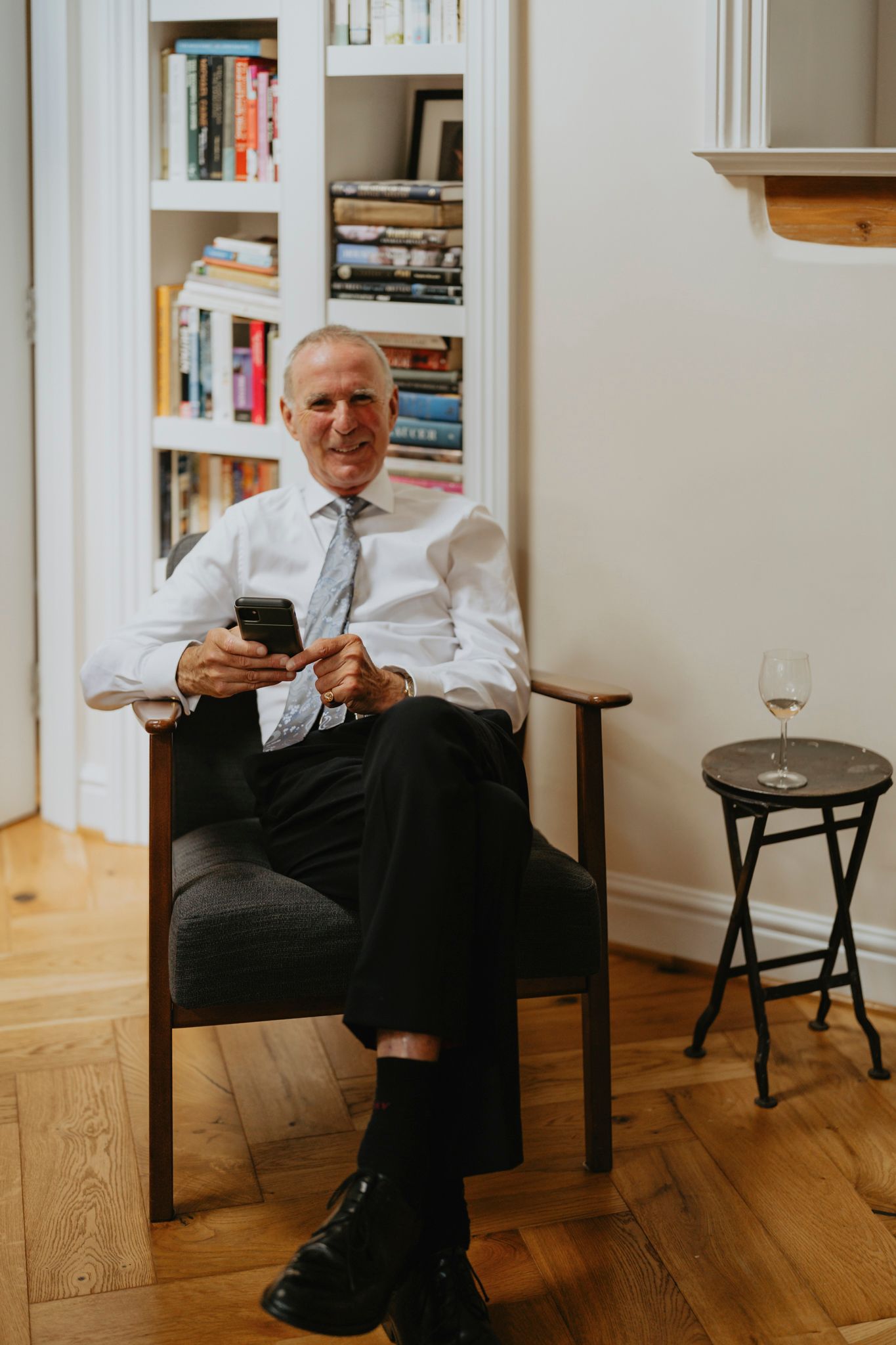 A man in business attire sits in an armchair checking his phone with a bookshelf and wine glass in the background.