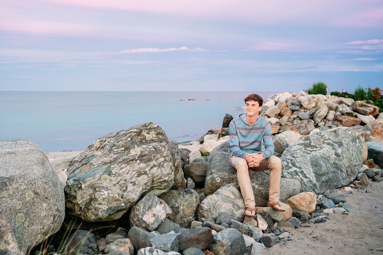 Someone sits with camera on rocky shoreline during sunset over calm waters and pink sky.