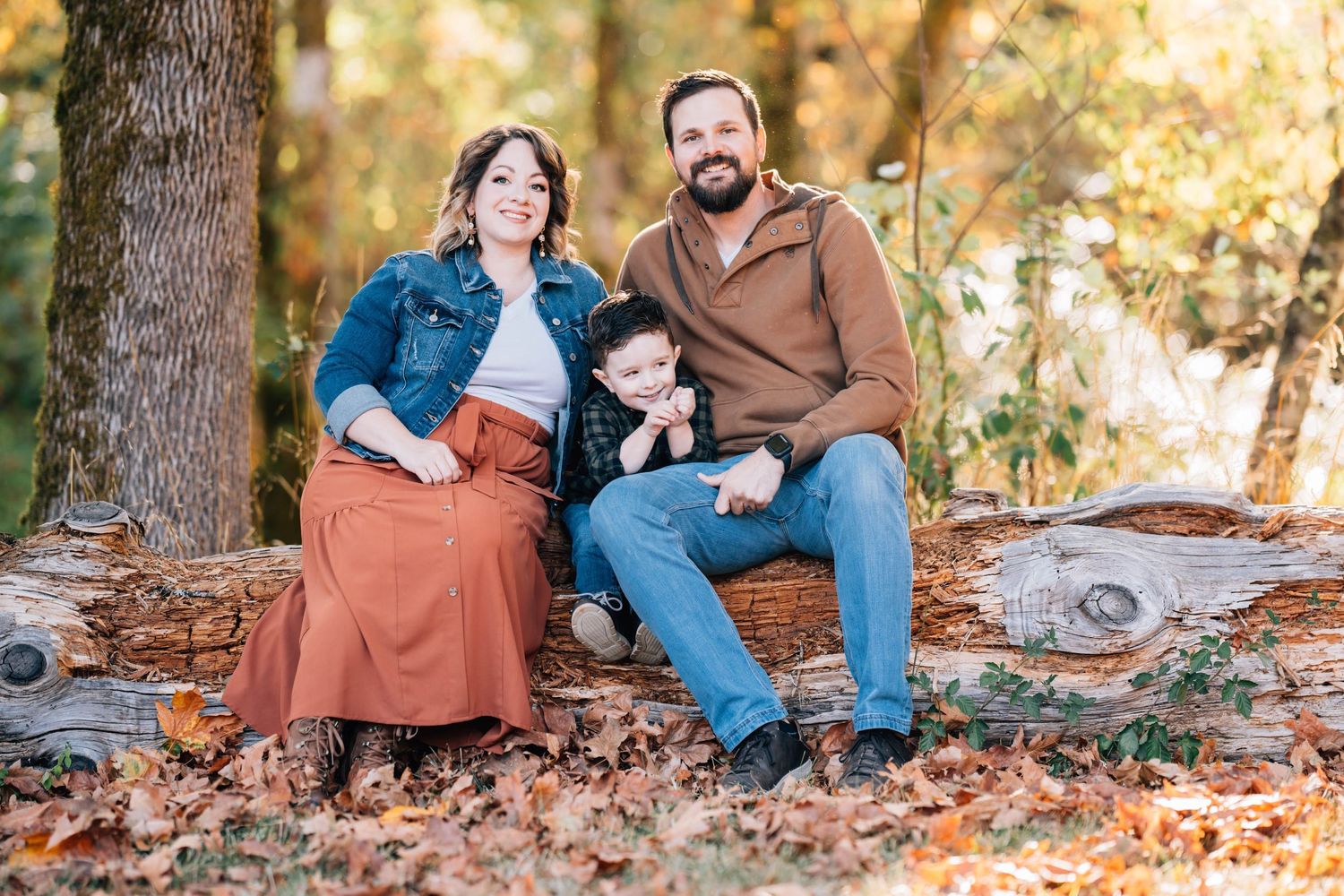 A small family sits together on fallen logs surrounded by autumn leaves in a wooded outdoor setting.