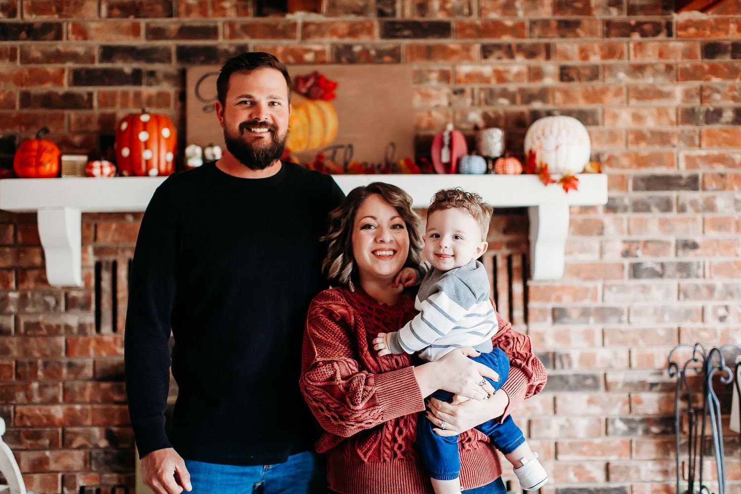 Three people pose for a family photo in front of a rustic brick wall with holiday decorations on a shelf behind them.