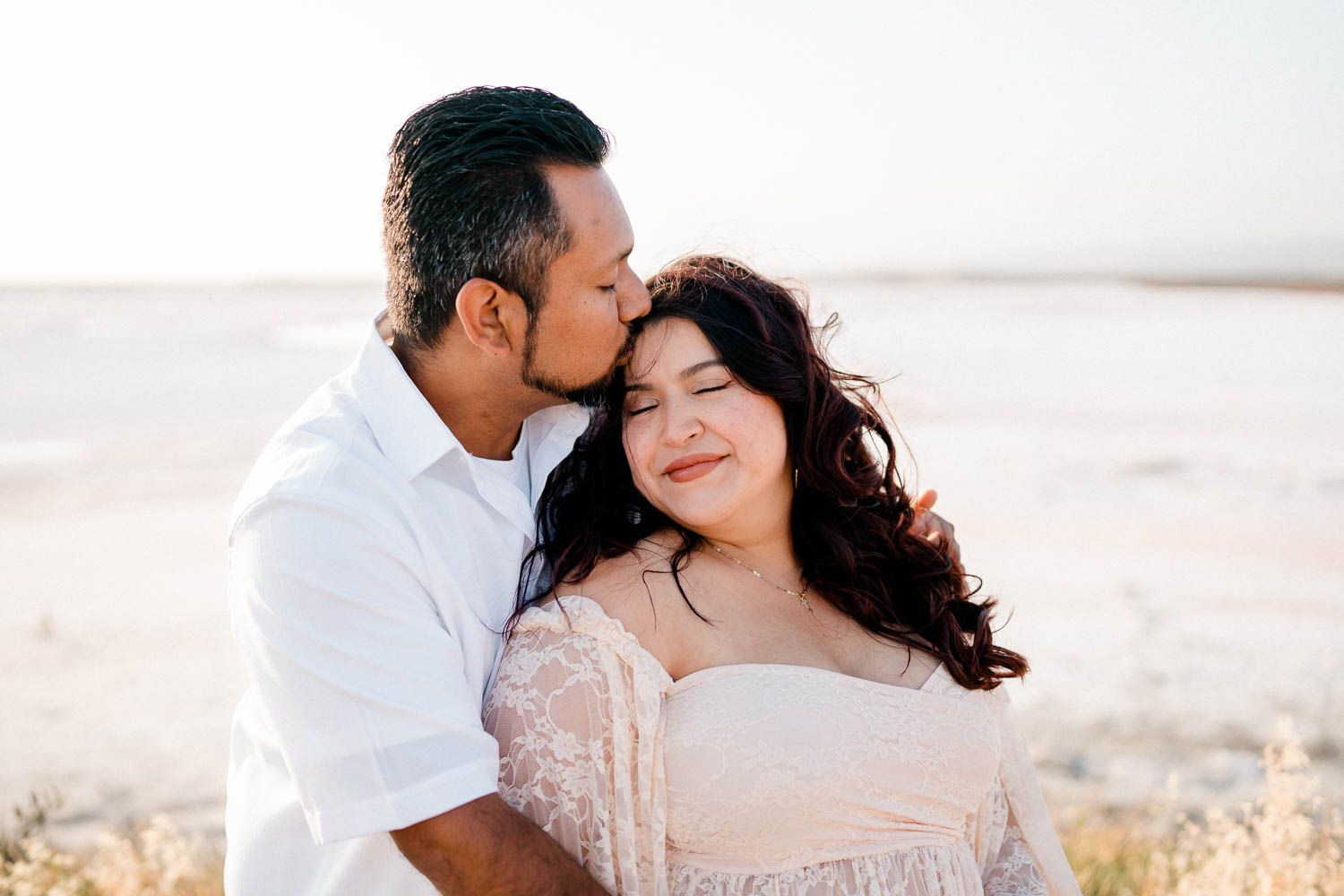 A romantic moment between a couple in white clothing standing together in a sun-drenched open field at golden hour.
