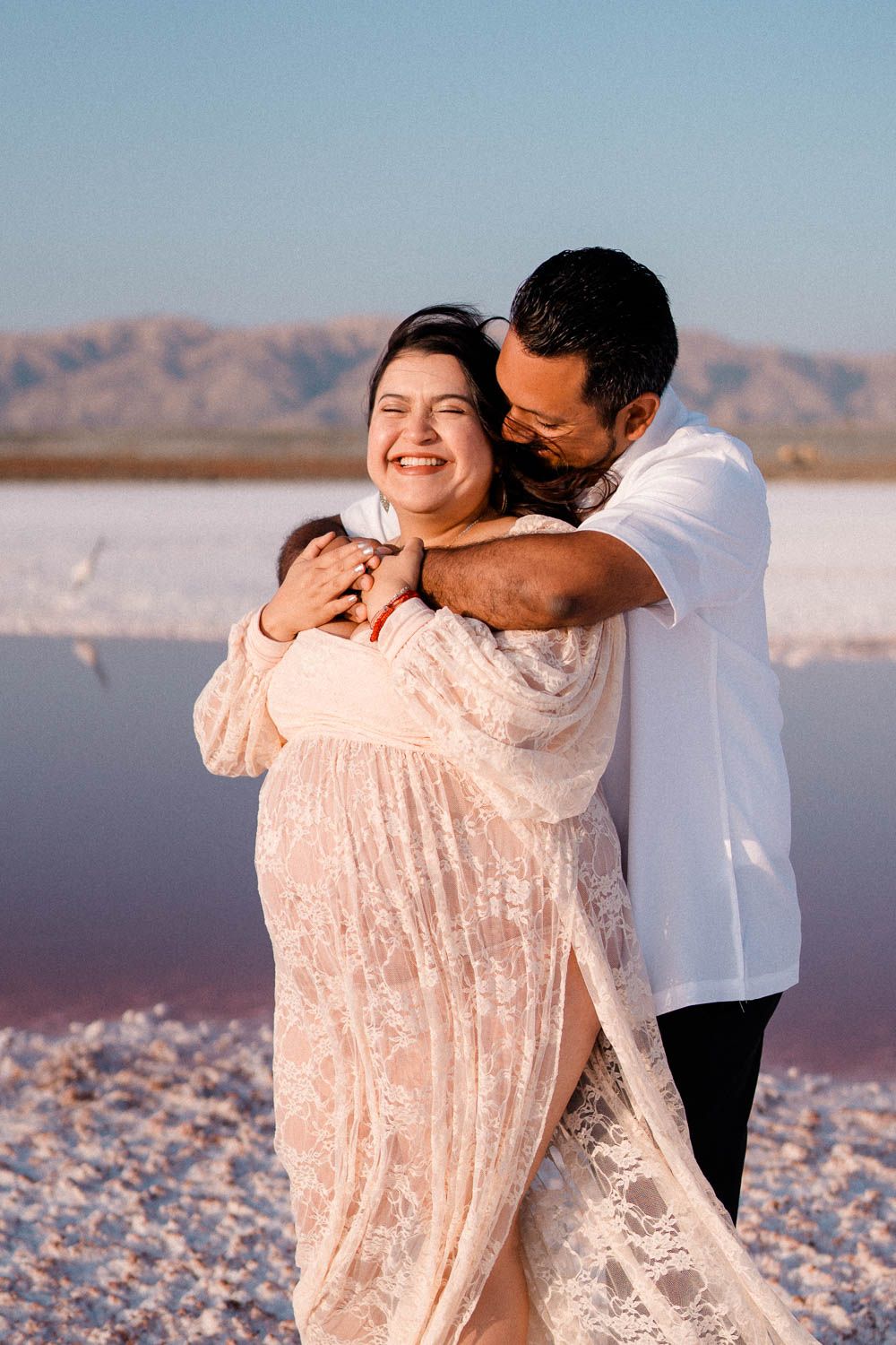 Couple embraces lovingly on salt flats at sunset wearing white and pink outfits against desert mountain backdrop.