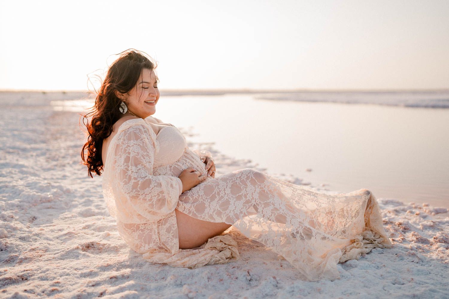 A pregnant woman in a white lace dress sits gracefully on a sandy beach at sunset.