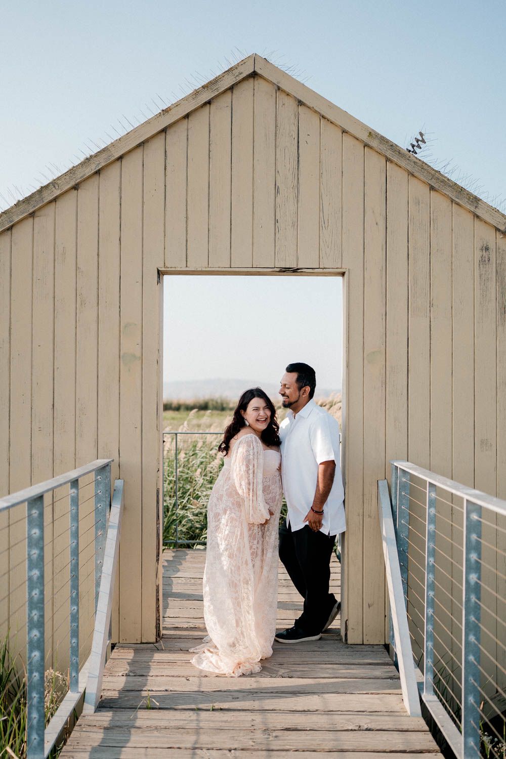 A couple poses in a wooden doorframe on a beachside boardwalk with light beige siding and metal railings.