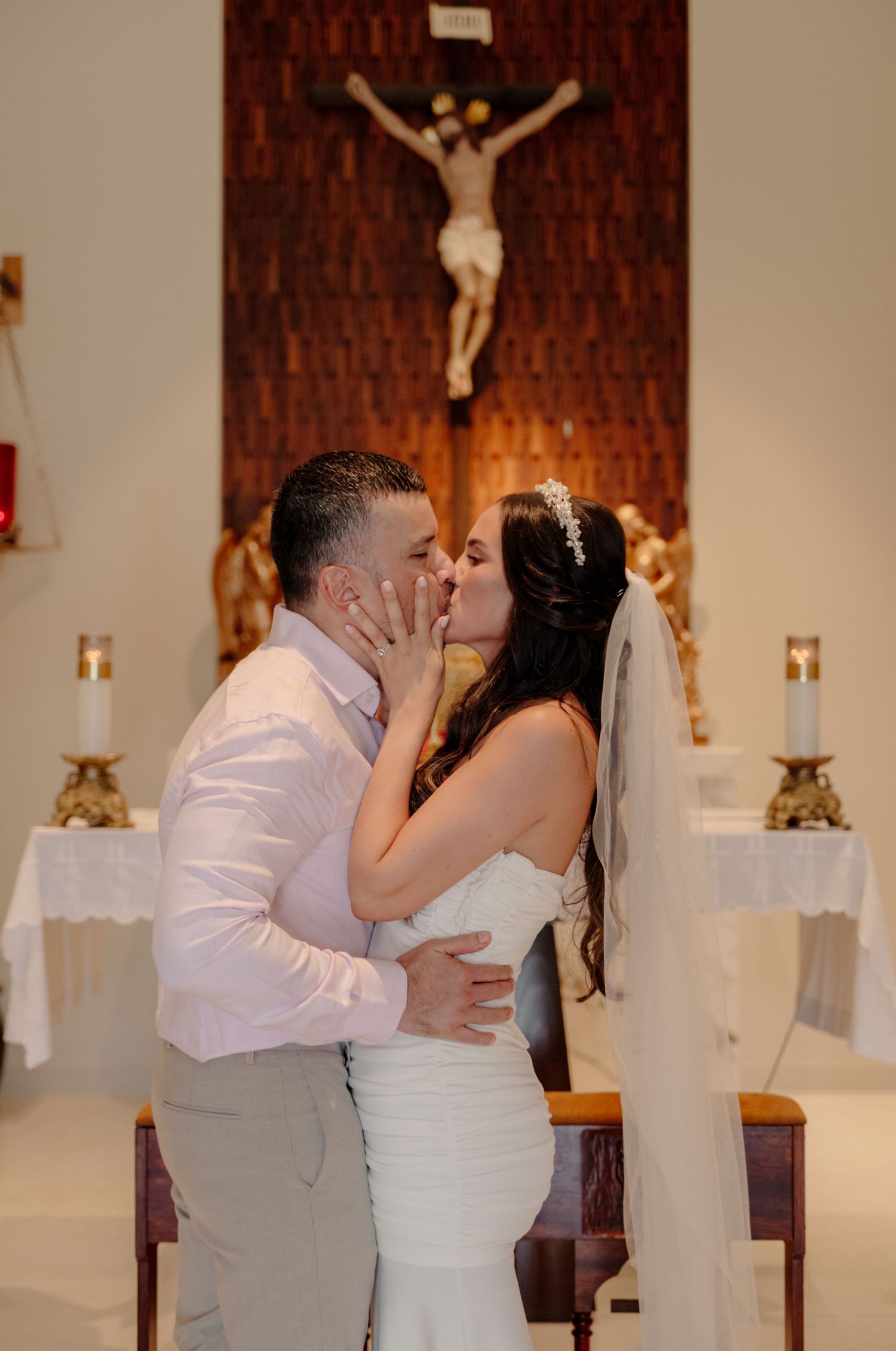 A couple shares their first kiss as newlyweds at the altar of a Catholic church with a crucifix on the wall behind them.