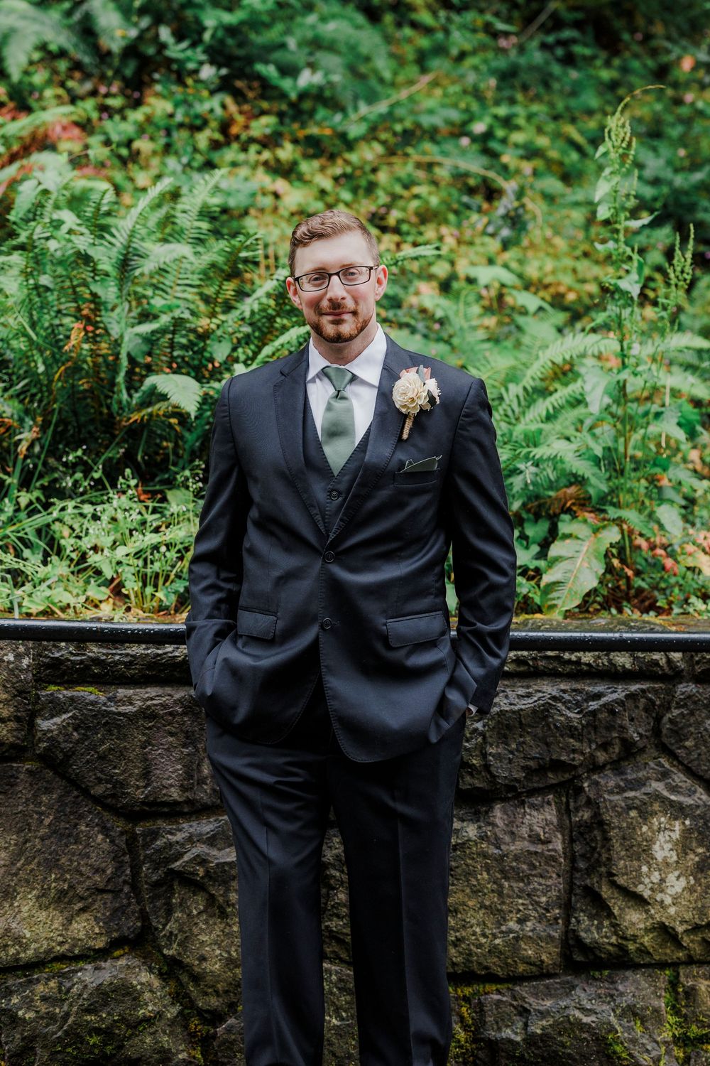 A formal wedding portrait of a groom in a black suit standing against a stone wall backdrop with lush greenery.