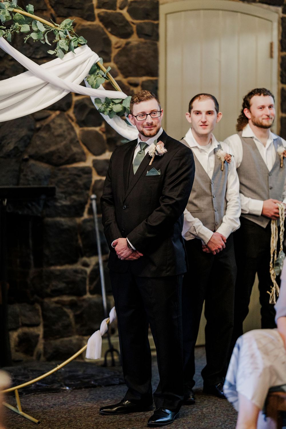 Groomsmen stand under a decorated arch with greenery and white fabric during a wedding ceremony.
