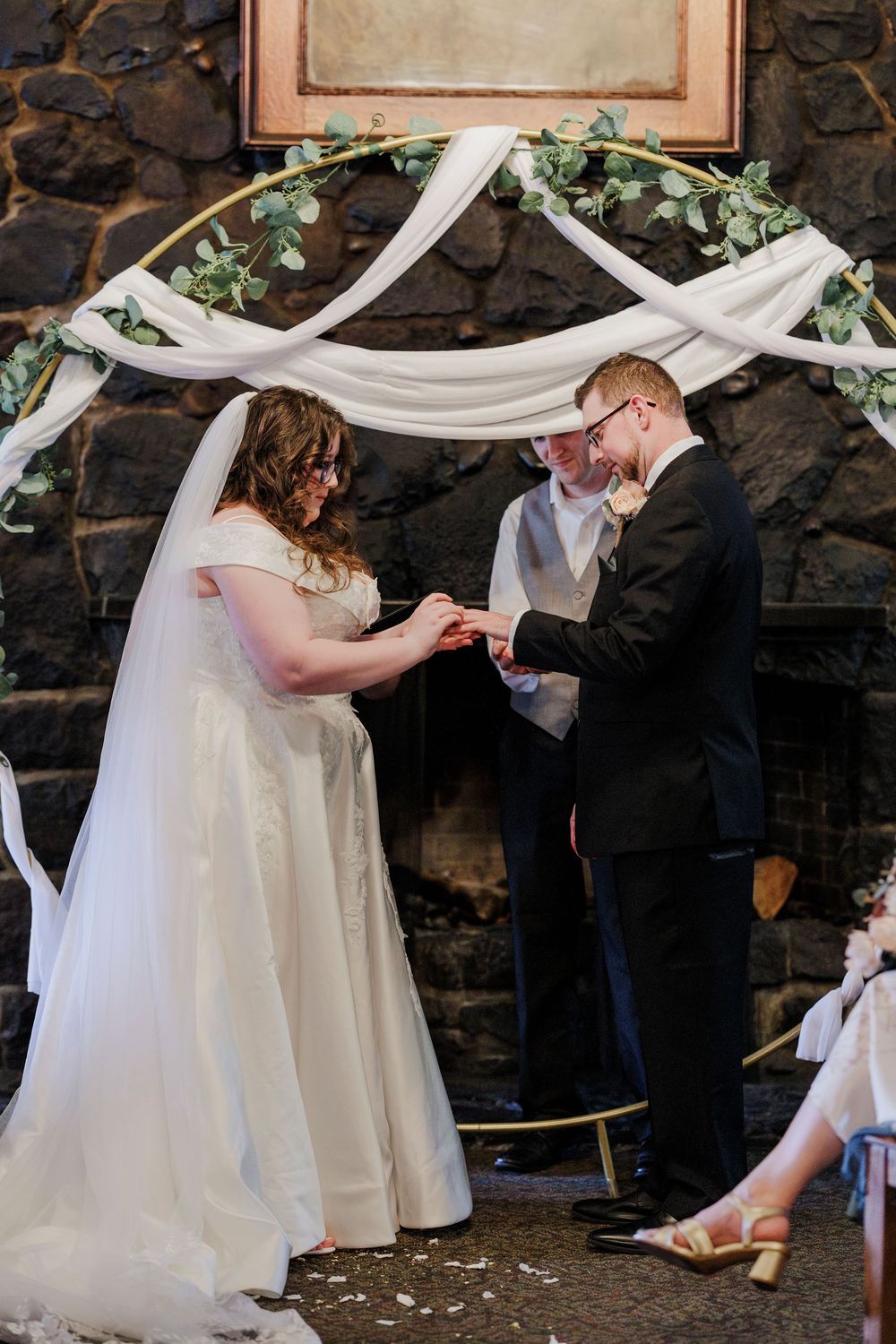 A couple exchanges rings under a white draped arch with greenery during their wedding ceremony.