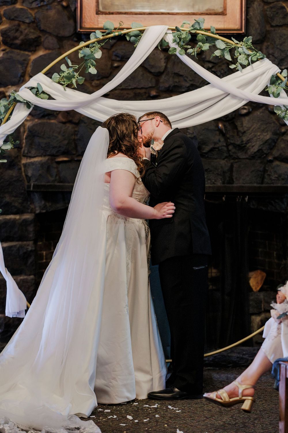 Wedding ceremony under white draped fabric and greenery arch against stone wall backdrop.