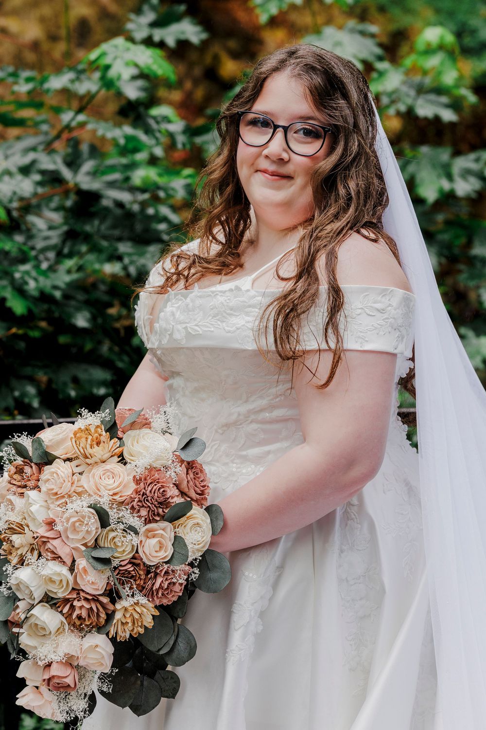 A bride in a white off-shoulder dress holds a peach and cream bouquet outdoors.