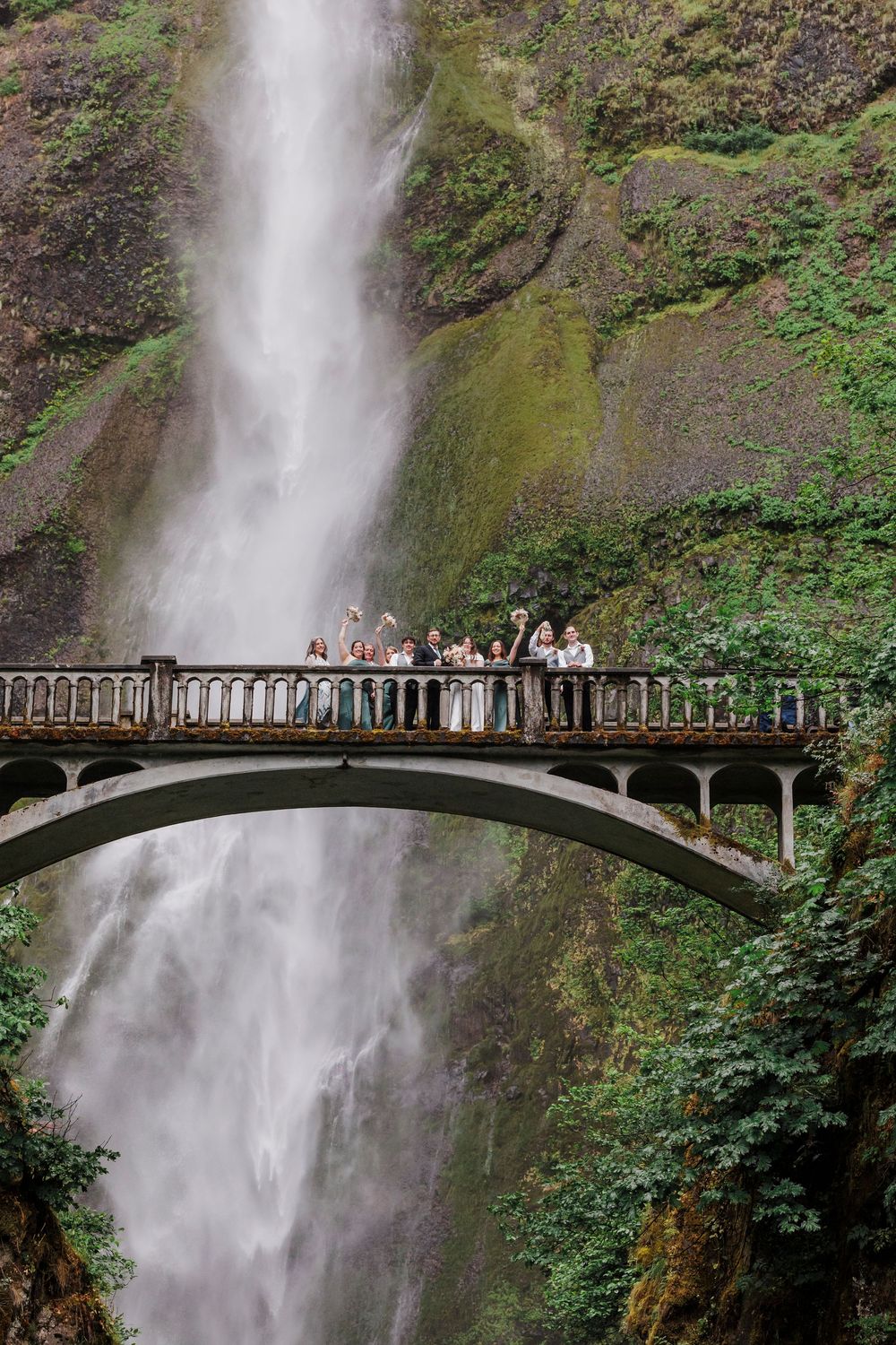 Wedding party gathered on scenic bridge overlooking dramatic Multnomah Falls waterfall in Oregon.