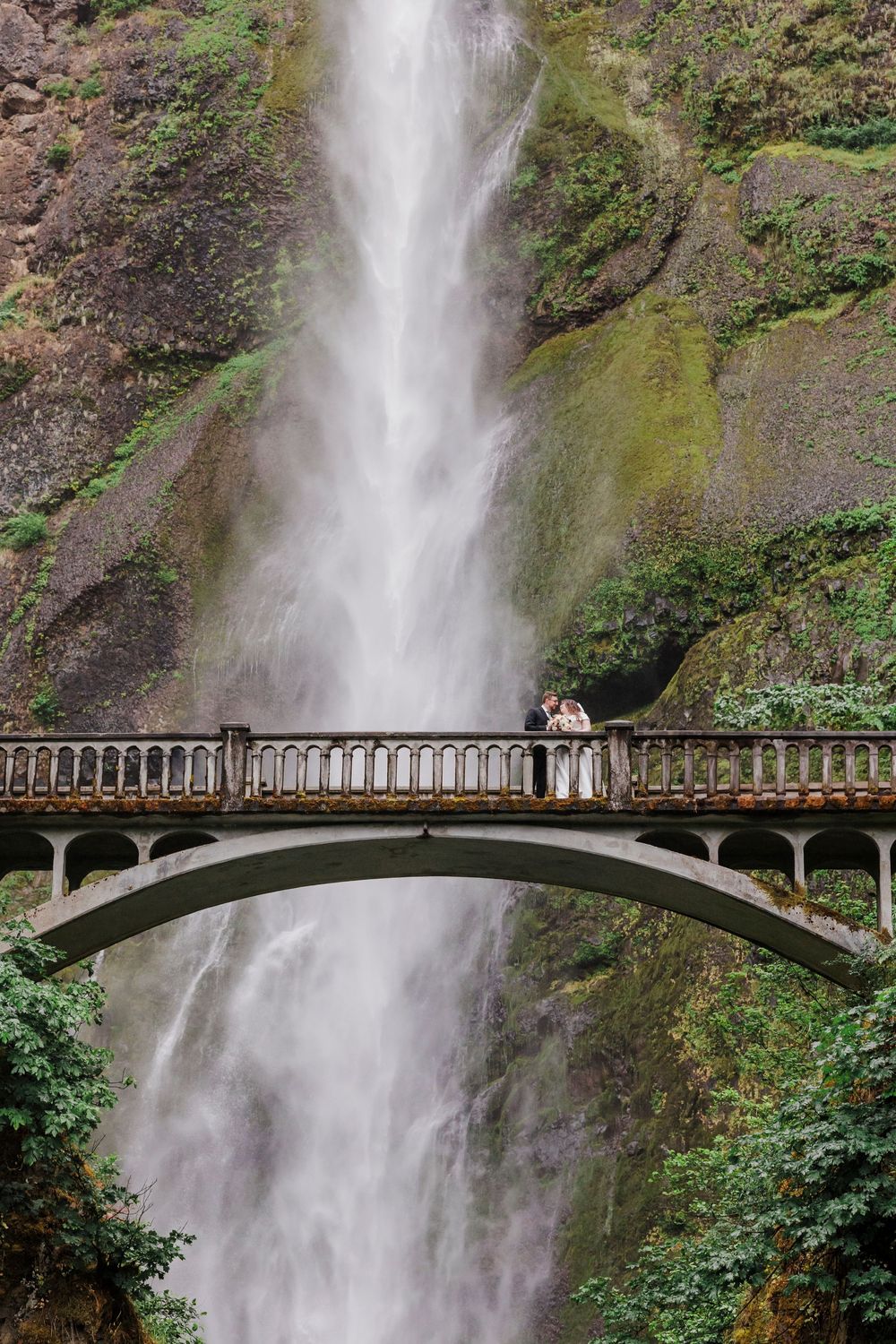 Stunning waterfall backdrop with a bridge crossing in front creating a dramatic wedding portrait setting.