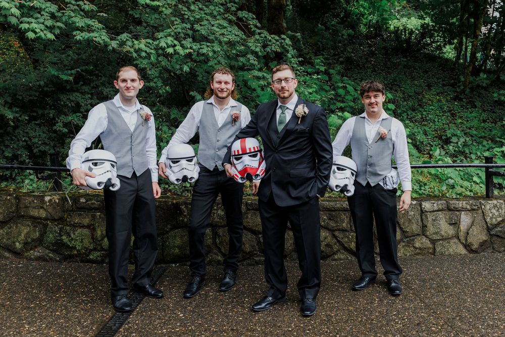 Groomsmen in formal attire pose holding Star Wars stormtrooper helmets outdoors.