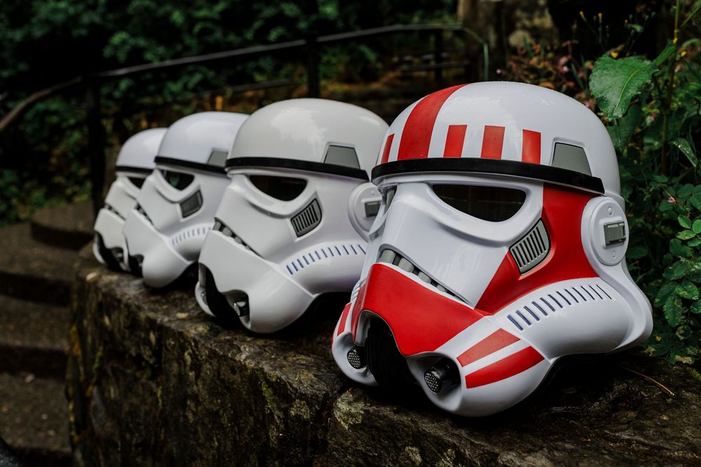 Two Star Wars Stormtrooper helmets, one with red stripes, displayed on a stone wall.