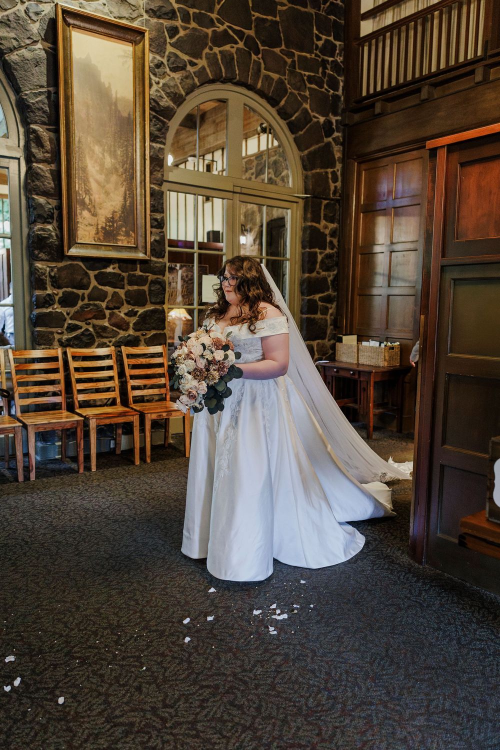 Bride holding bouquet stands in elegant stone-walled venue with arched windows and wooden details.