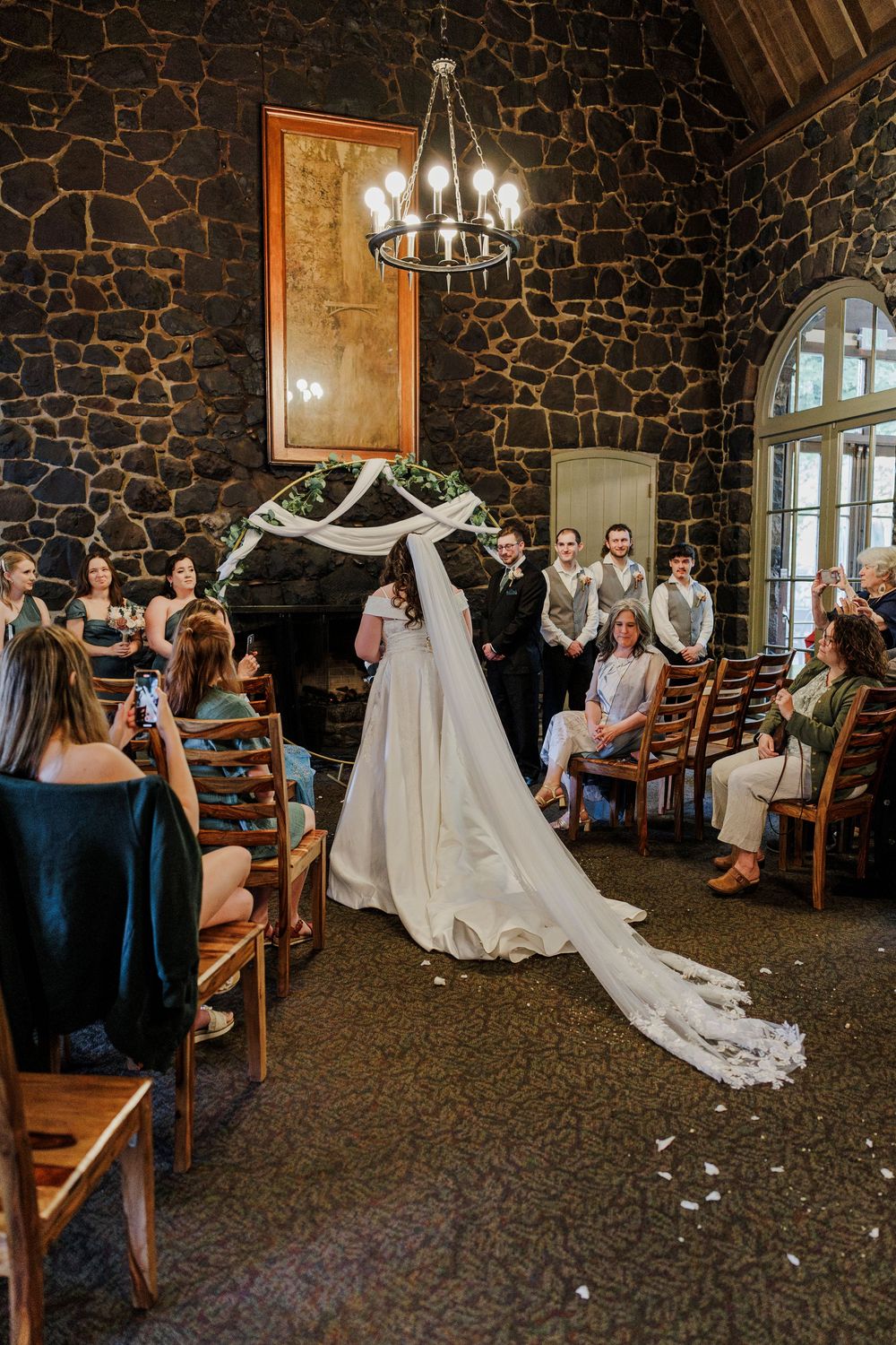 Wedding ceremony in progress inside rustic stone chapel with chandelier and wooden chairs.