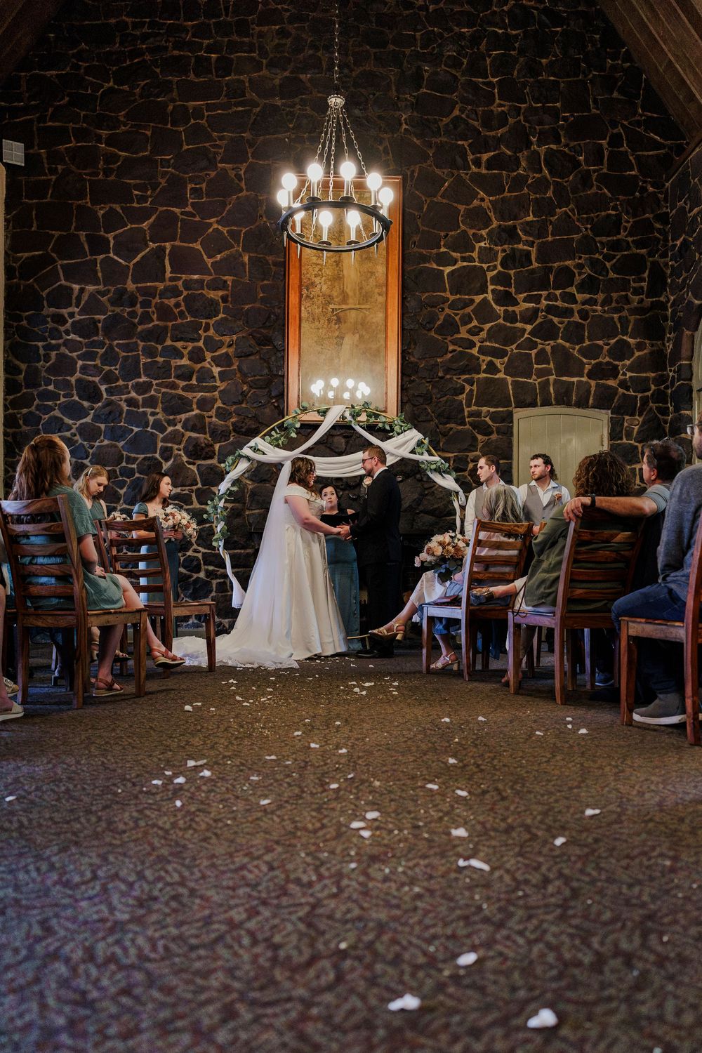 Wedding ceremony in rustic stone-walled venue with chandelier and scattered flower petals.