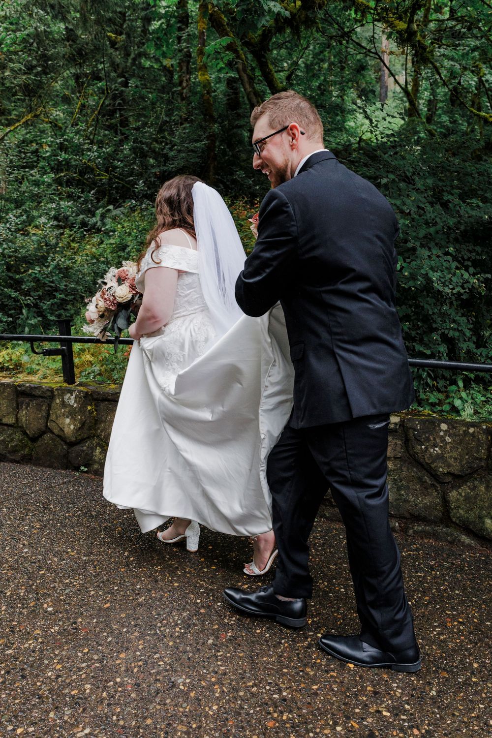 A wedding dress being adjusted and fluffed outdoors on a stone path surrounded by green foliage.