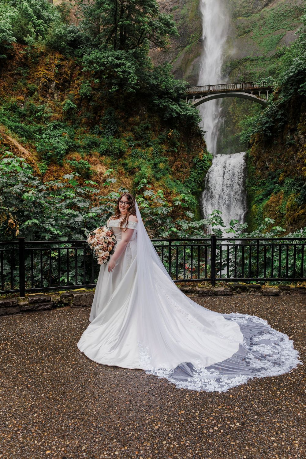 Bride in flowing white gown poses before Multnomah Falls with dramatic train and bouquet.