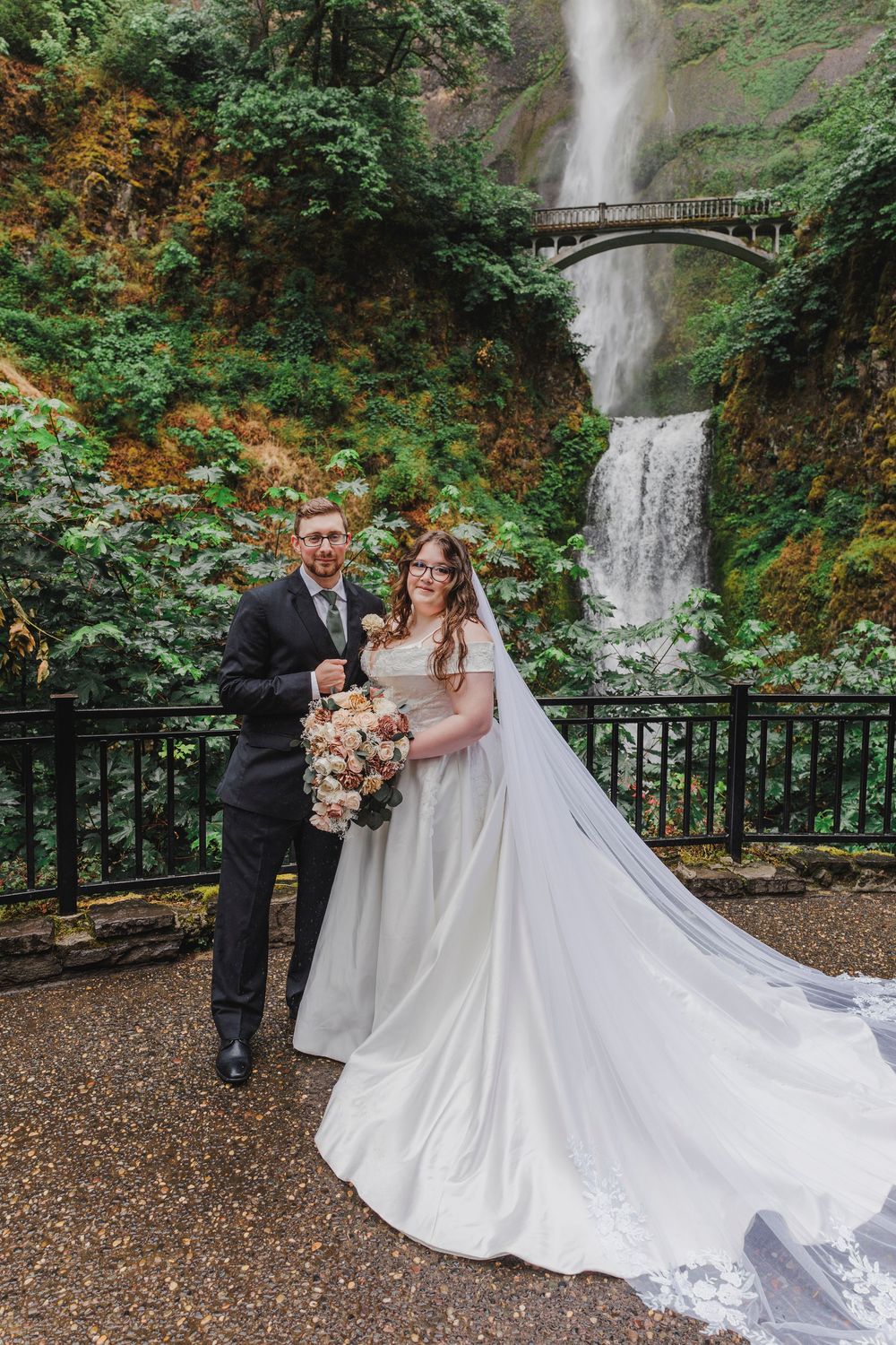 A wedding portrait at Multnomah Falls in Oregon with a dramatic waterfall and stone bridge in the background.