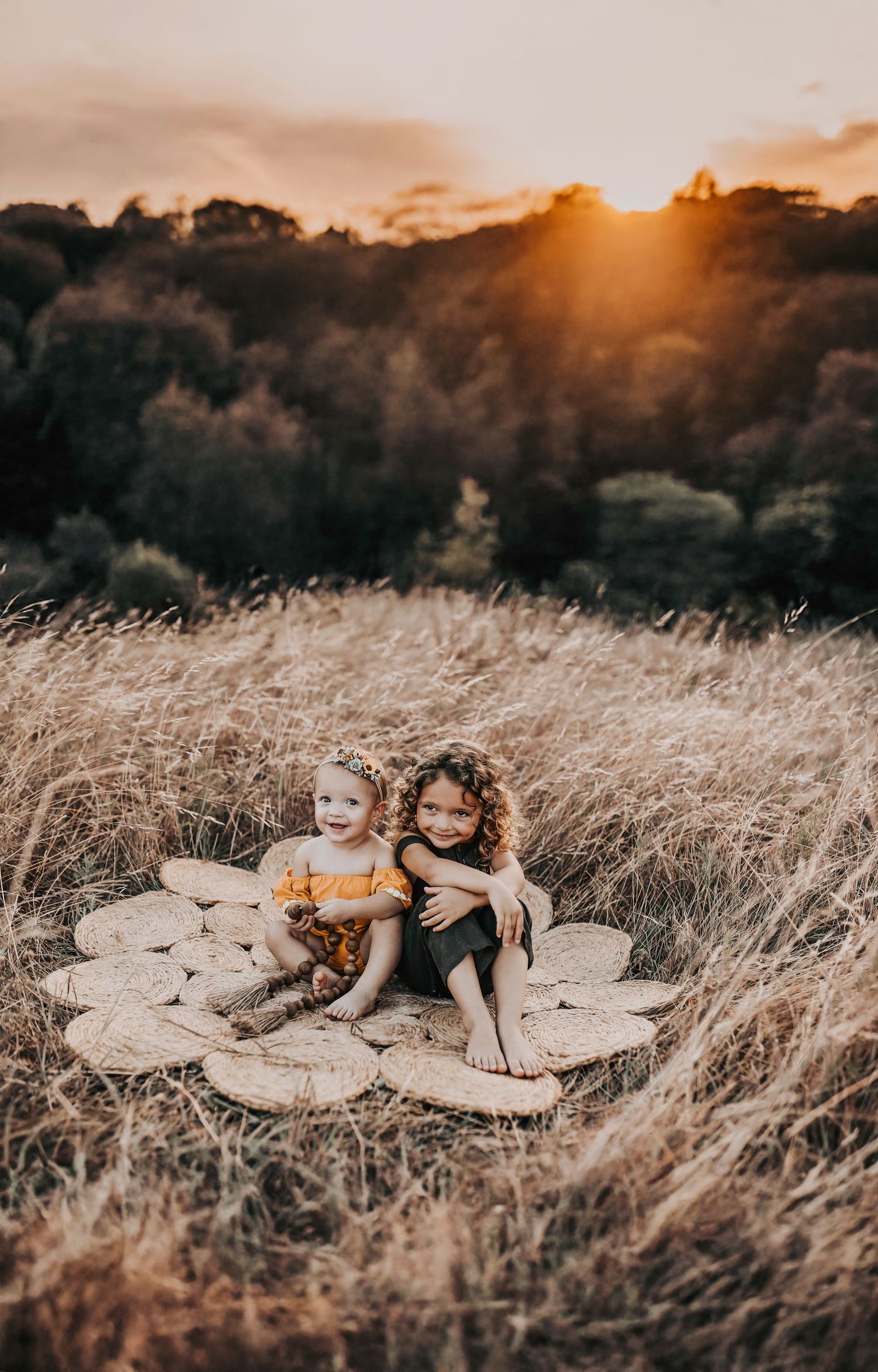 sisters snuggle while playing on a natural fiber rug in the middle of a field of golden grass