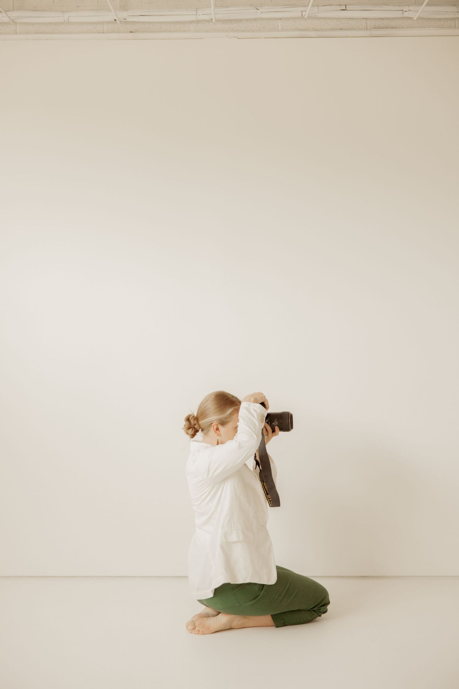 Person in white shirt and green pants kneeling on floor while taking photo with vintage camera against light background.