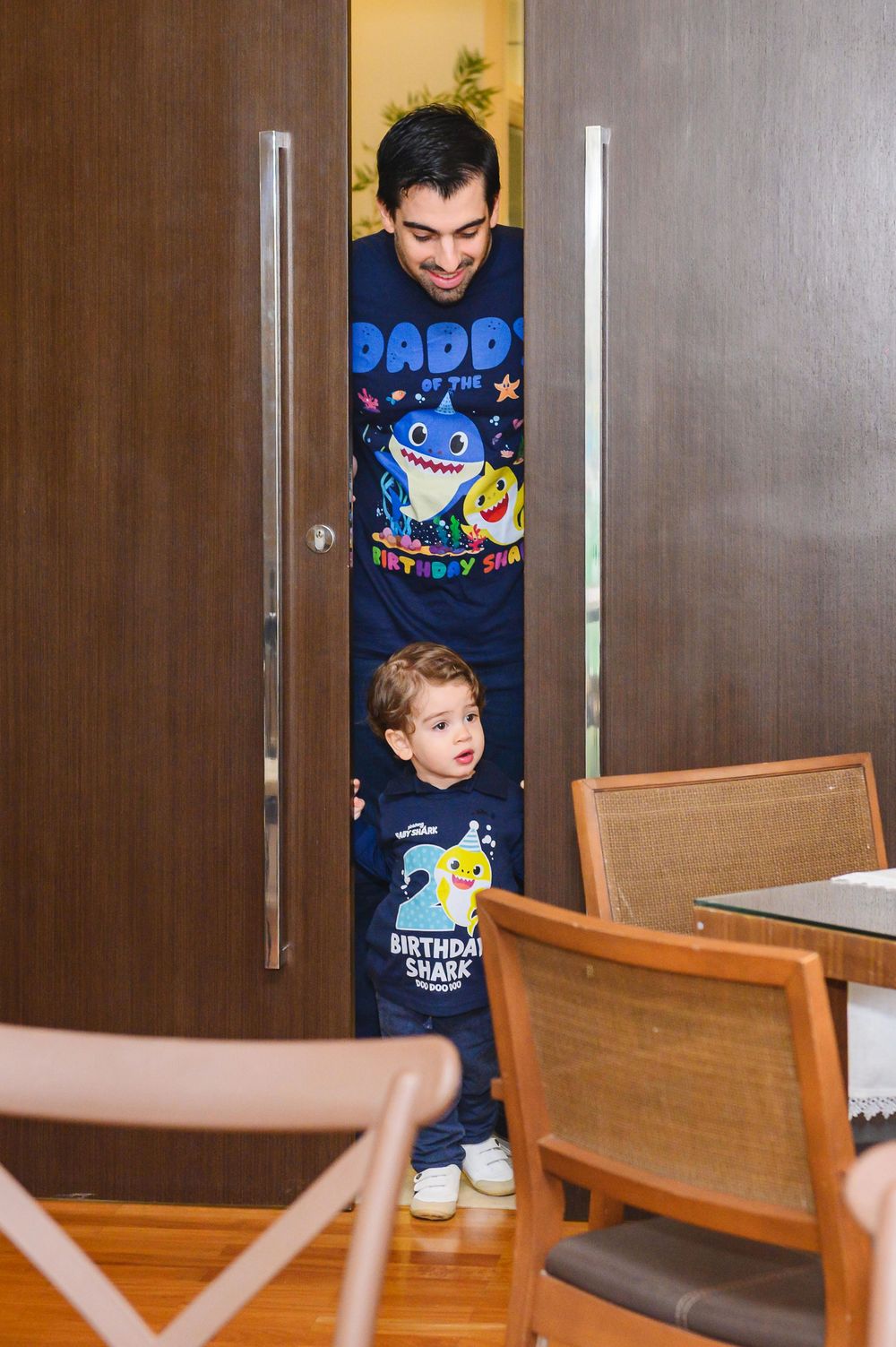 Two people peek playfully from behind a wall in what appears to be a dining area with wooden chairs.