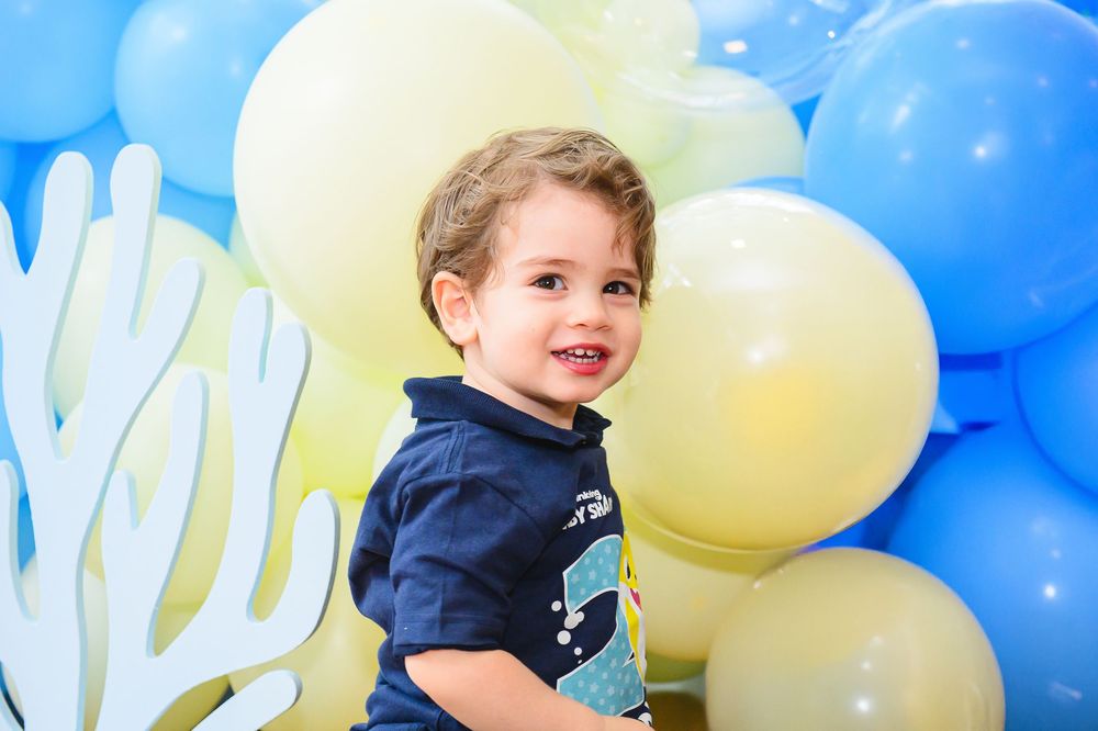 Young child in navy blue shirt smiling in front of a colorful balloon backdrop with blue and yellow balloons.