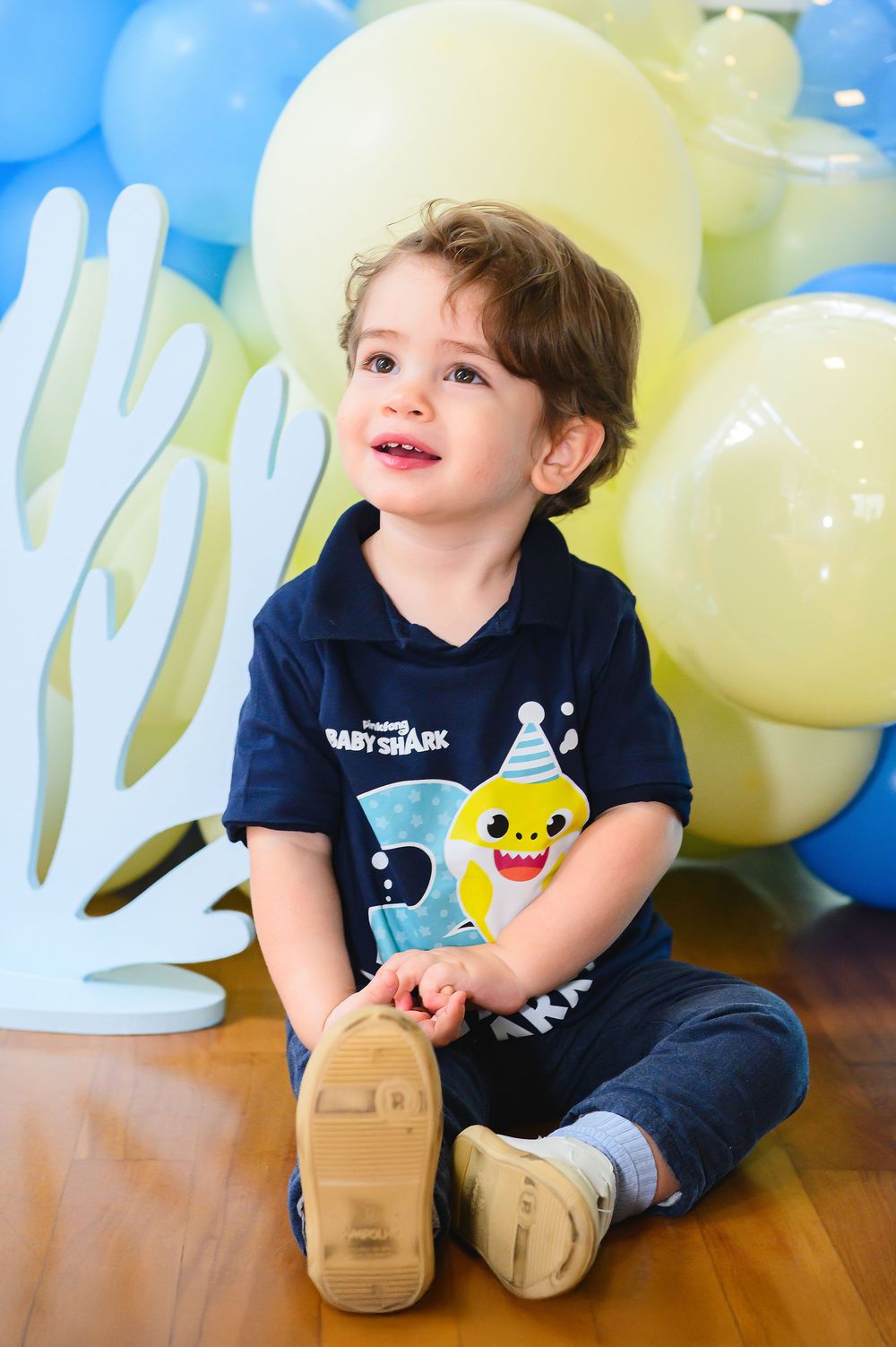A young child sits on wooden flooring wearing a Baby Shark navy blue shirt surrounded by blue and yellow balloons.
